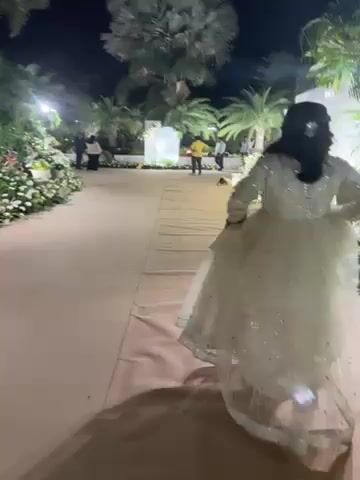 At night, a woman in a sparkly white dress is walking on a beige carpet pathway that is lined with white flower arrangements. There are black lights along the pathway. In the background, other people are visible, including two men in yellow shirts talking. There are palm trees and other greenery in the background, and some white arches are visible. The video is slightly shaky and filmed from behind the woman walking.