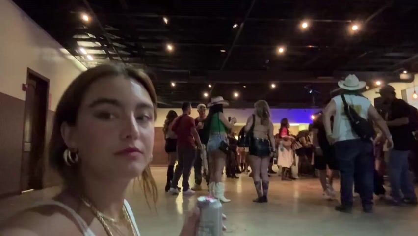 A young woman with brown hair and hoop earrings, wearing a white top and a gold chain, looks towards the camera and speaks, holding a silver can. Her expression is lively. Behind her, a diverse crowd of people mingle in a dimly lit indoor venue. Many individuals in the background are dressed in casual attire, with some sporting cowboy hats and boots, suggesting a country or western-themed event. The space features exposed ceiling structures, illuminated by warm string lights and some distant purple ambient lighting. The floor appears to be a light-colored, possibly concrete surface. The scene portrays a social gathering or concert environment during the evening or night.