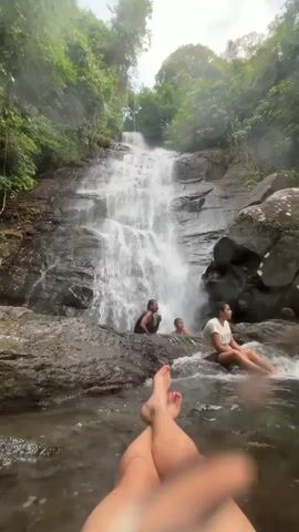 The video shows a rocky waterfall in a lush, green environment. There are three people in the middle ground of the shot, two of whom are standing in the water at the base of the waterfall and one is sitting on a rock on the water’s edge. The viewer’s legs and feet are in the foreground.