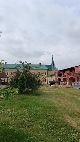 The video shows a panoramic view of a religious complex under a cloudy sky. In the foreground, there is green grass with white flowers and a small tree with green leaves. Behind the grass, there is a red building with white trim and arched windows. A portion of a green-roofed building with a tall green spire is visible in the background. The video pans from left to right, revealing more of the red building's facade and a pathway lined with bushes. There are no people or animals visible in the video.