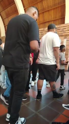 The video shows a gathering of people indoors, under a wooden-paneled roof. The floor is tiled with alternating light and dark squares. The people are of varying ages and are dressed in casual clothing, including jeans, t-shirts, and hoodies. Some are wearing hats or sunglasses. Several individuals are seen walking around, suggesting the atmosphere is relaxed and social. The presence of a girl in a pink top and a boy in a pink hoodie indicates the event may be family-oriented. The overall scene appears to be a casual gathering, possibly a party or public event, taking place indoors.