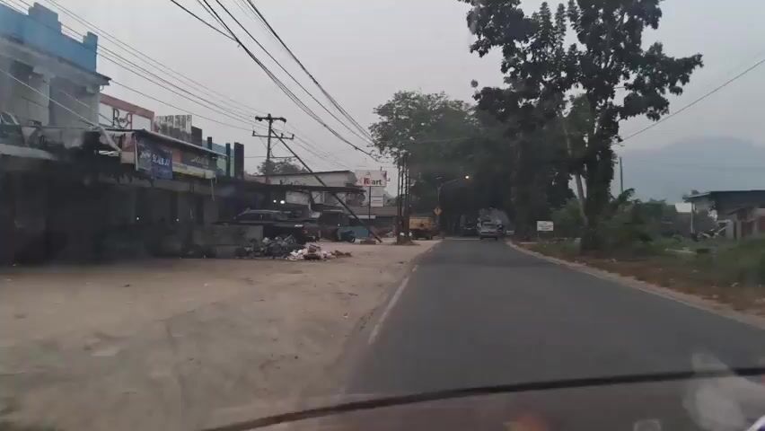 The video begins with a view of a street scene on an overcast day. On the left, there is a row of low-rise buildings with awnings and signs, one of which appears to be for "R Mart." There is debris and trash piled on the ground in front of these buildings. Several vehicles, including what appear to be pickup trucks and motorcycles, are parked or visible in front of the buildings. Overhead, numerous power lines are strung across the scene. To the right of the street, there is a paved road with several cars and a yellow truck traveling away from the camera. Tall trees with dense foliage line the right side of the road, and a hazy mountain range is visible in the distance. The overall impression is of a slightly disorganized, developing area.