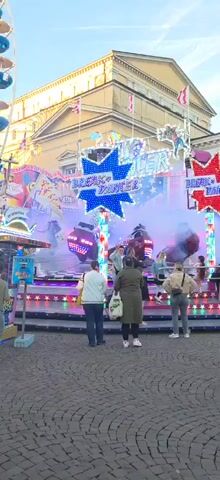 This video captures a vibrant scene at an amusement fair in Darmstadt, Germany. The central subject is a "Break Dancer" ride, brightly illuminated with numerous colorful LED lights in shades of blue, green, red, and yellow. Large star-shaped signs on the ride prominently display the text "BREAK DANCER" in blue and red. Another section of the ride structure shows the word "CASINO."

The ride is actively in motion, with several individuals seated in its spinning cars, which oscillate and rotate, creating an exhilarating experience. A white, hazy mist or smoke effect adds to the dynamic atmosphere around the ride. Many people are seen standing on the cobblestone ground surrounding the ride, either watching the action, waiting to board, or simply enjoying the fair environment.

In the background, a large, light-colored building with classical architectural elements, possibly a theatre or historic hall, stands under a clear, bright sky, suggesting late afternoon or early evening. A tall Ferris wheel with blue and yellow cabins is partially visible on the left side of the frame, indicating other attractions at the fair. The camera pans and slightly rotates, capturing the energy and movement of the scene. The overall impression is one of festive entertainment and enjoyment at an outdoor event.