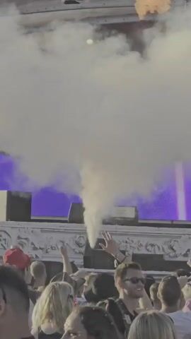 This video depicts a large outdoor music festival crowd gathered in front of a stage. The subjects are primarily people enjoying the event, with some objects like speakers and lighting rigs visible on and around the stage. The scene is characterized by a hazy, smoky atmosphere, likely from a smoke machine, which partially obscures the stage and the sky. The overall activity is a performance at a festival, with the audience engaged and looking towards the stage.

Notable details include the time of day appearing to be daytime, given the bright lighting. The weather seems clear and sunny, as indicated by the brightness and the clear sky in some parts of the video. The location is suggested by the text "AIRBEAT ONE" prominently displayed on the stage structure, a known electronic dance music festival held in Neustadt-Glewe, Germany. The emotions of the people in the crowd appear to be excitement and enjoyment, with many raising their hands in the air. The presence of a large, elaborate stage structure further emphasizes the scale of the event.
