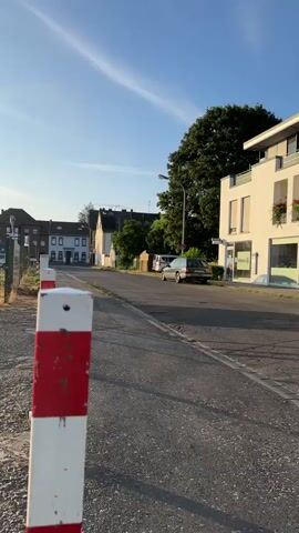 A red and white striped pole is in the foreground of the video. The camera pans up and to the right. The scene then shifts to a street with houses and trees. There is a car parked on the side of the street, and another car drives past. The sky is blue with a few wispy clouds. The sun is shining, and the lighting suggests it is either early morning or late afternoon.