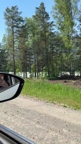 A car is traveling on a dirt road, with the rearview mirror visible in the foreground. Beyond the road, there is a dirt embankment with grass and trees. In the distance, there is a body of water and a forest. An excavator is seen operating on the embankment, digging into the dirt. A white car is parked on the road further down. The sky is clear and blue.