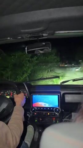 The video shows the interior of a car at night, with the driver's hands on the steering wheel. The dashboard is illuminated, and a large screen in the center displays a colorful interface. The headlights of the car are on, illuminating a dirt road ahead with some vegetation on the sides. In the distance, other vehicles with their headlights on can be seen. The camera is positioned from the passenger's seat, looking forward. There is some motion blur, indicating the car is moving.