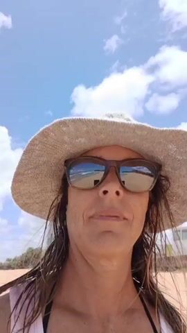 A woman in a white tank top, a light-colored hat, and sunglasses is filming herself on a sandy beach. The beach and a building with blue and white paint can be seen in the background. The sky is mostly blue with some puffy white clouds. The woman is talking. The reflection of the beach and sky is visible in her sunglasses.