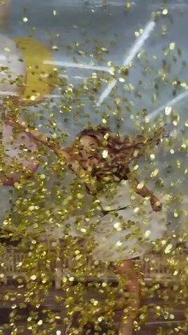 A young woman with blonde, curly hair is captured mid-motion, her body angled as if jumping or running. She wears a white dress with black trim and is holding a cluster of three balloons, one pink and two yellow, in her left hand. Golden confetti is falling and swirling around her, obscuring parts of her body and the background. The background appears to be a photographic mural depicting a cityscape with a bright sky and white lines, possibly representing lights or infrastructure. The overall impression is one of celebration or joy.