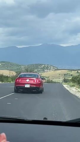 A red Ford Mustang, seen from the rear, drives along an asphalt road. The car has a Bulgarian license plate, PB TB 0538. The road curves slightly to the right. The scenery includes rolling hills with sparse trees and vegetation. In the distance, a mountain range is visible under an overcast sky. A second car is seen approaching in the distance. The video is recorded from the perspective of another car following the Mustang. The dashboard is visible in the foreground.