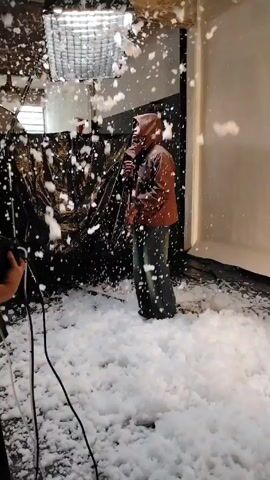 The video shows a person standing in a room, covered in artificial snow. The person is wearing a hooded brown jacket and dark pants. The floor is covered in a thick layer of artificial snow, and more snow is falling from above. There's a lighting fixture above the person, and what appears to be a backdrop behind them. Someone is holding a device that is spraying the snow. The scene appears to be a studio setup for a photoshoot or filming. A light stand and cables are also visible.
