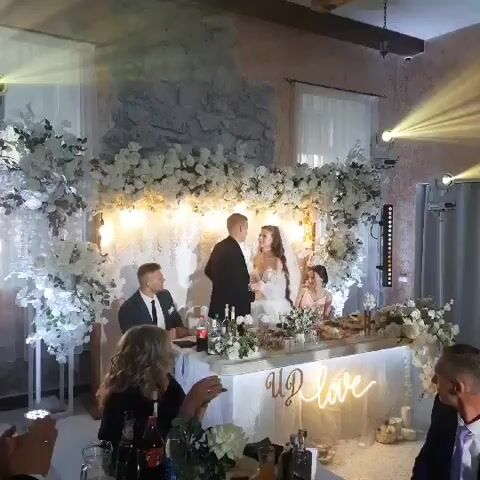 The video shows a wedding reception with a bride and groom kissing at a decorated head table. The table is adorned with white flowers, candles, and a neon sign that reads "Up love." A man in a suit sits to the left of the couple, and a woman in a pink dress sits to the right. The room is lit by spotlights and string lights. In the foreground, several guests are visible, including a woman clapping her hands and a man looking at the camera.