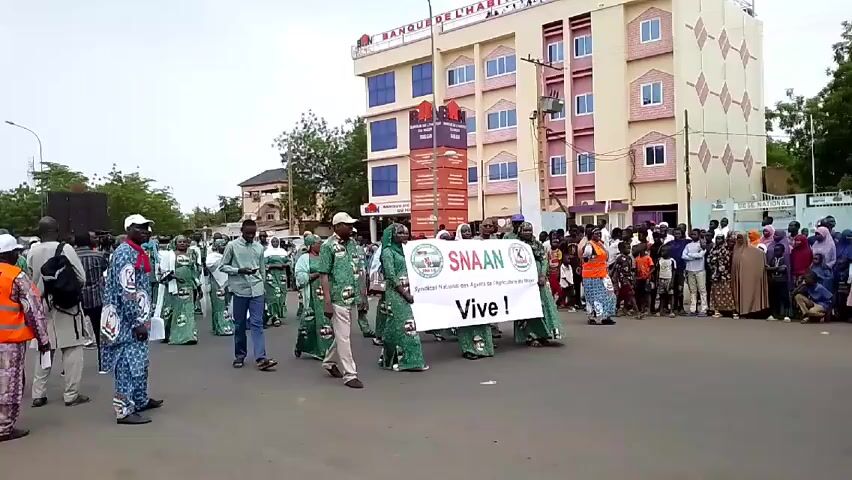 The video captures a procession of people walking along a street, likely in an urban area in an African country. Most participants appear to be wearing matching green and blue patterned outfits, with some wearing white caps. They are holding a large banner with "SNAAN" and the phrase "Vive!" prominently displayed, along with some smaller text. A crowd is lined up along the sides of the street, appearing to watch the procession. The background includes a multi-story building with windows and signage, power lines, and some trees. The overall environment suggests a daytime event, potentially a parade or demonstration, in a setting that seems to be either a city center or a well-developed suburban area.