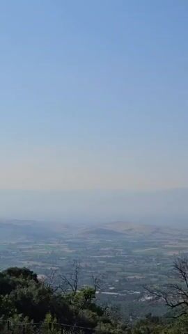 A video records a slow, steady rightward pan across a wide, elevated view of a landscape under a clear blue sky. The initial frame displays a sprawling city nestled in a valley, flanked by distant, hazy mountains. In the foreground, dark green treetops are visible. As the camera pans right, more of the city and valley floor become visible. Further into the pan, a cluster of houses with various architectural styles, including modern designs and traditional red-tiled roofs, appears on the hillside in the foreground, interspersed with dense green trees. The pan concludes with a prominent white minaret with a blue pointed roof appearing on the far right, standing tall above the trees and houses on the hillside, with the distant city and mountains still visible further left in the background. No people are visible.