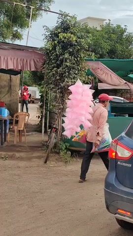The video shows an outdoor scene at what appears to be a roadside stall or market. In the foreground, a dark blue car is partially visible on the right. To the left of the car, a person in a light-colored shirt and a red cap is standing next to a stall covered with a green tarp. The person is holding a large, pink, fluffy object, which resembles cotton candy. Behind this person and the stall, a tall, leafy tree stands prominently. To the left of the tree, a bicycle is leaning against something. Further left, there is a small structure with a pink and white awning. Inside this structure, a person in a red jacket and a red cap is visible, holding what looks like a toy or a similar item. Two chairs are placed outside this structure. The ground is dirt and gravel. The sky is overcast.