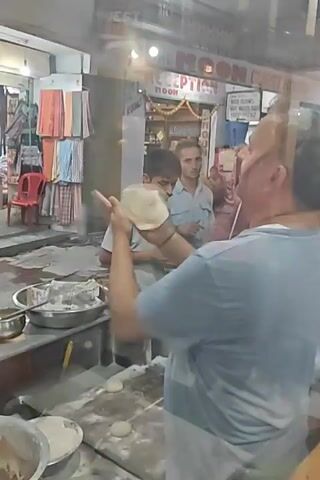 The video captures a lively street scene, likely at an open-fronted food stall or restaurant in Kasol, India, during the evening or night, indicated by the artificial lighting. In the foreground, a man in a light blue t-shirt is actively preparing flatbreads (possibly naan or roti). He is seen flattening pieces of dough on a floured surface before skillfully placing them into a cylindrical tandoor oven.

Beside him, a younger man in a light grey t-shirt stands near a counter, holding a stack of what appears to be freshly baked flatbreads in a woven basket. Bowls containing dough and flour are visible on the counter, along with other kitchen implements.

In the background, several people are walking or standing on the street. A prominent sign for "THE MOON GUEST HOUSE" and "RECEPTION MOON" is visible, suggesting the food stall might be part of or near a guesthouse. Other text like "ROOMS" can also be seen. The setting suggests a bustling market or tourist area with various shops and establishments. The overall atmosphere appears casual and busy, with the focus on the food preparation process.