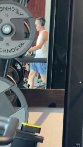 A man in a white tank top and blue shorts with headphones on is walking on a treadmill in a gym. The camera is focused on weight plates on a machine in the foreground, with the man visible in a mirror behind the plates. The man has a tattoo on his upper left arm. The gym appears to have wood-paneled walls.