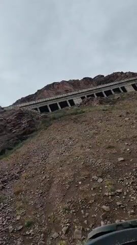 The video shows a red truck in the foreground, tilted at an angle. In the background, there is a rocky, mountainous landscape with a large, weathered, building structure built into the hillside. The sky is overcast. The camera appears to be moving, possibly from inside the truck.