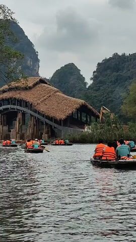 The video shows several boats on a river. Each boat carries several people wearing orange life vests. The people are rowing their boats in the river. In the background, there is a bridge with a thatched roof. The bridge is surrounded by mountains and trees. The sky is overcast.