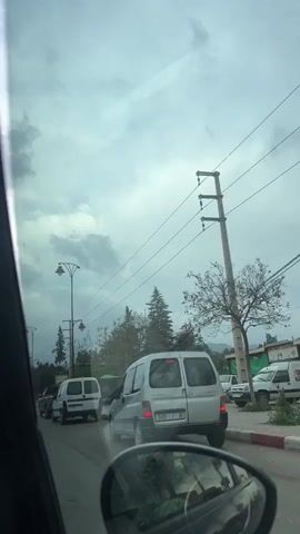The video captures a street scene in Ourika, Morocco, from the perspective of a moving vehicle on a cloudy day. The sky is overcast with light grey clouds throughout the footage.

Several vehicles are visible in traffic, including a grey Peugeot Partner van with a roof rack on the right, displaying the license plate "9491 I 36." Another white van, with "CITR" (likely Citroën) partially visible, and a black pickup truck with "TOYOTA" clearly written on its tailgate, are also part of the traffic flow. A dark SUV is seen moving on the left.

The street features typical infrastructure: tall, decorative street lights with multiple lamps, numerous power lines strung between utility poles, and curbs painted in red and white stripes. Along the road, various trees can be seen, including evergreen-like species and palm trees, indicating a mix of vegetation. A small, green corrugated metal shack is visible on the right side of the street.

Pedestrians are observed walking on the sidewalks. Notably, an elderly man dressed in white clothing and a straw hat, using a cane, walks along the pavement. A sign with Arabic text, though unreadable, is also present. The overall scene depicts a bustling road in a town or village setting under an overcast sky.