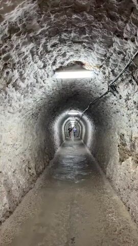 The video shows a perspective from inside a long, dimly lit, rough-hewn tunnel. The walls and ceiling of the tunnel appear to be made of rock or salt, with a rough, uneven texture. Fluorescent lights are mounted at intervals along the ceiling, providing the primary illumination. The floor of the tunnel is wet and appears to be made of concrete or compacted earth. Two individuals, a younger person in a dark t-shirt and shorts and an older person in a blue shirt and jeans, are walking away from the camera towards the end of the tunnel. Further down the tunnel, other figures can be seen in the distance, also walking. The tunnel extends into the distance, appearing to curve slightly.