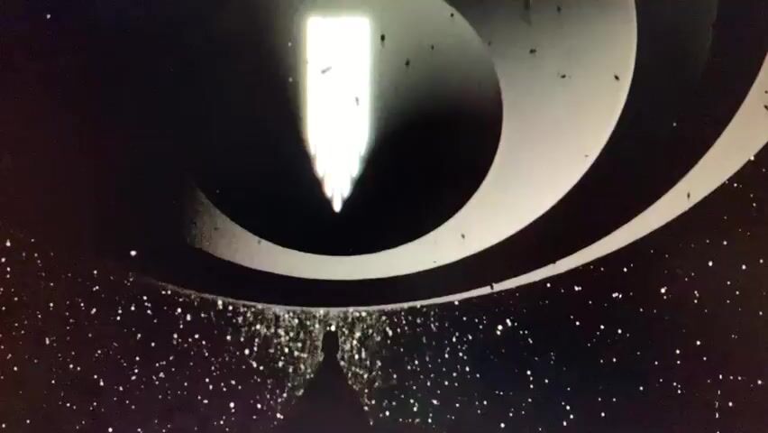 The media file depicts a stylized, abstract opening sequence. The dominant visual elements are a black background with a swirling, circular, metallic-looking structure that appears to be a tunnel or a vortex. At the top of the vortex, a bright, vertical white light emanates from a rectangular shape with vertical lines. At the bottom of the vortex, a lone, silhouetted figure stands on a triangular platform, facing the light. The background is filled with what appears to be a scattering of white particles, resembling stars or dust.

The activity is a visual representation of an entrance or a journey. The figure at the bottom seems to be either approaching or observing the intense light at the top, which suggests a destination or a revelation. The swirling vortex creates a sense of motion and depth, drawing the viewer's eye towards the central light source.

There are no specific indications of time of day or weather. The setting is abstract and non-realistic, evoking a feeling of space, technology, or perhaps a metaphorical journey. The emotion conveyed is one of mystery, awe, and possibly anticipation due to the contrast between the dark surroundings and the bright, central light.

There is visible text in the form of a logo or title within the vertical white light at the top of the vortex. The letters are stylized and white against the light, but difficult to discern clearly due to the resolution and stylization. The letters appear to form a word or name, but are not legible enough to be definitively identified.

The provided location context of "Pisochyn, Ukraine" does not appear to be directly represented in the visual content of the media file, which is abstract and symbolic.