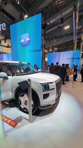 The video shows a white Zeekr 9x vehicle on display at what appears to be an auto show. The car's doors are open, revealing its interior. The background features a large blue screen displaying the text "A I everything A I verywhere" and an abstract icon. People are visible around the car, likely attendees of the show, observing the vehicle and interacting with it. The environment is well-lit and spacious, typical of a convention or exhibition hall. The camera angle slightly moves, focusing primarily on the car’s exterior and partially capturing the surrounding show environment.