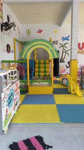 The video shows an indoor playground area with colorful walls decorated with cartoon animals and hot air balloons. In the center is a large yellow and green play structure with a slide and a ball pit. To the right of the play structure, there is a yellow seesaw and another slide. The floor is covered with blue and yellow interlocking foam mats. Several children are present, playing in the playground. One child, wearing a blue shirt and grey pants, is seen sliding down a slide. Another child, wearing a blue shirt and black pants, is playing with toys on the floor. The room appears to be well-lit and clean.