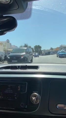 The video is shot from the interior of a car, looking forward through the windshield. The car is in motion, driving down a street lined with buildings and trees. Sunlight streams through the windshield, causing some glare. On the dashboard, a car stereo system is visible. The street appears to be in a sunny, possibly Mediterranean, location. Several other vehicles are on the road, including vans, cars, and a motorcycle parked on the side. A person wearing a red shirt and white pants is walking across the street in the distance.
