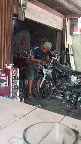 A man wearing a dark blue t-shirt, grey shorts, and a baseball cap is working on a black motorcycle. He is standing and appears to be tightening something on the front of the motorcycle with a tool. The motorcycle is partially disassembled, with parts of the engine and frame visible. To the left of the man, there is a red object, possibly another motorcycle, and a speaker system. Various cans and bottles are scattered around the workspace. The scene appears to be an outdoor workshop or garage with a corrugated metal roof and a wooden structure. In the foreground, a glass is visible on a table.