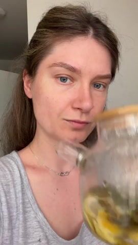 A young woman with light brown hair, tied back partially, and blue eyes is shown up close. She is wearing a grey tank top and a delicate silver necklace with a double infinity pendant. The woman is in the process of drinking from a clear glass teapot that contains sliced lemons and possibly mint leaves. The teapot has a light-colored lid, possibly bamboo. The background is a plain, light-colored wall, suggesting an indoor setting, likely a home.

The activity captured is the woman drinking from the teapot. Her eyes are closed as she takes a sip, and then they open as she pulls away from the spout, with a slight smile and a contented expression. The lighting is bright and even, suggesting it is daytime. There are no visible external cues indicating the time of day or weather. The location is specified as Ożarów Mazowiecki, Poland, but this context does not provide specific visual information within the frame.

There is no visible text within the media file. The focus is entirely on the woman and the teapot, highlighting a moment of refreshment.