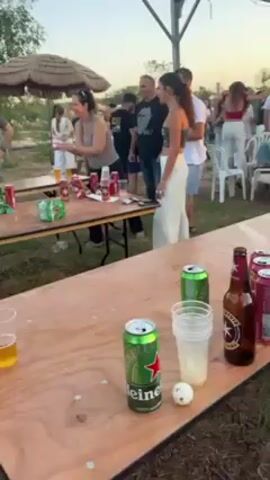 The video shows a group of people playing beer pong outdoors. There are two long wooden tables set up with cups of beer, beer cans, and ping pong balls. Some people are gathered around the tables, drinking and watching. One woman is throwing a ball towards the cups. There's a thatched umbrella in the background and a barbecue grill. The atmosphere seems casual and social.