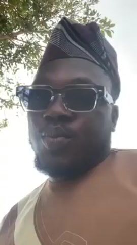 A close-up shot shows a man outdoors, wearing a traditional patterned hat and sunglasses. He has a beard and is wearing a brown and beige patterned top. He is looking at the camera with a neutral expression. Behind him, there are trees with bare branches against a bright sky. In the background, a tent and some people can be seen. The camera angle is slightly tilted upwards, looking at the man.