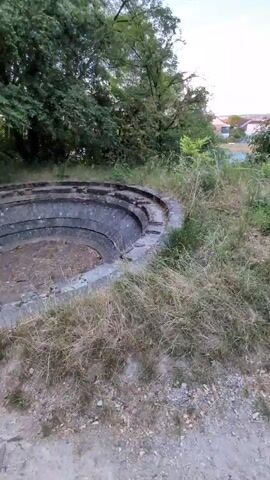 The video shows a circular, tiered concrete structure partially overgrown with dry grass and weeds. The structure appears to be outdoors, with trees and foliage visible in the background. The concrete is worn and weathered, and the central area is filled with dirt and debris. The lighting suggests it is daytime, possibly late afternoon or early evening. There are no people or animals visible in the video. The camera is positioned at ground level and slowly pans around the structure.