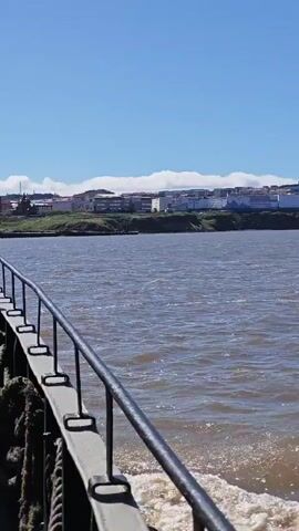 The video is taken from the perspective of a boat moving through choppy, brown water. The camera pans across a wide expanse of water, with a shoreline visible in the distance. The shoreline features a town or city with buildings and some industrial structures, including cranes. The sky is clear and blue with a few wispy clouds. A railing and rope are visible in the foreground, indicating the viewer is on deck. The water churns behind the boat, creating a wake.