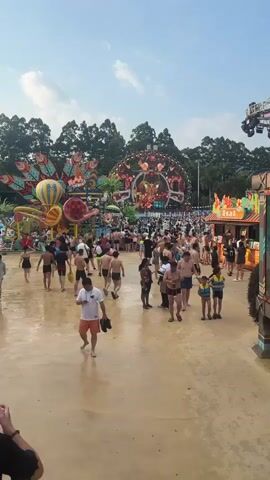 The video shows a busy outdoor water park with many people milling around. In the background, there is a large, colorful stage with lights and decorations, suggesting a performance or event. Several water slides and other attractions are visible, adding to the festive atmosphere. People are dressed in swimwear, and many are walking, talking, or standing in groups. The ground appears to be wet, indicating it's a functioning water park. The sky is partly cloudy, suggesting it is daytime.