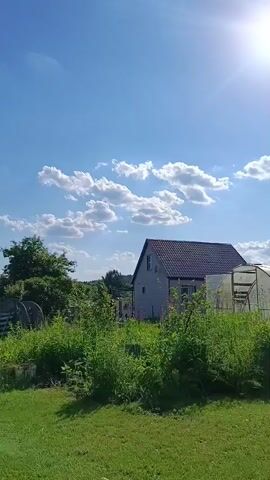 The video opens with a shot of the sky and some clouds, then pans to show a yard with a house, a greenhouse, and various plants. A person's feet are visible in the foreground, resting on the grass. The camera then moves to reveal a white hanging chair, followed by a woman standing at a table in the yard, cutting something. The scene appears to be outdoors on a sunny day.