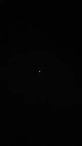 The video depicts a small, distant light source against a black background. As the video progresses, the light source appears to change color intermittently, flickering between white, red, and green. There are no other discernible objects or details in the video.