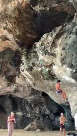 A man is rock climbing on a cliff face, secured by a rope. There are three people standing on the beach below, watching him. One person, a woman, is holding a phone and appears to be filming or taking a picture. The environment consists of a sandy beach and a large rocky cliff face. The climber is wearing shorts and climbing shoes, and the people on the beach are dressed casually. The scene takes place in an outdoor setting with natural lighting.