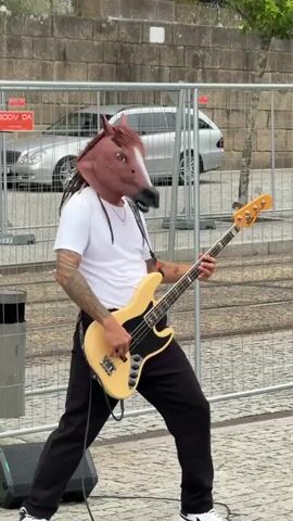 A person wearing a horse mask, black pants, and a white t-shirt plays a cream-colored electric bass guitar. They are standing in front of a stone wall and a metal fence. Behind the fence, a silver car is parked. The person's left arm has tattoos. The bass guitar has four strings. A black speaker is visible on the lower left of the frame. The ground is paved with cobblestones.