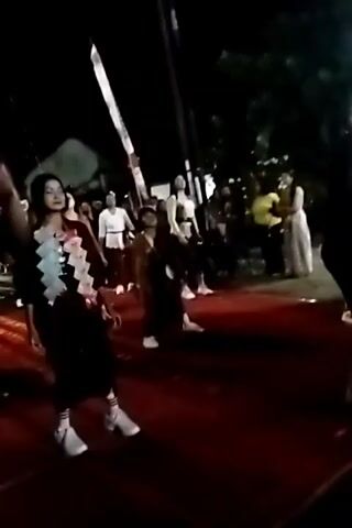 The video begins with a close-up of a person wearing a dark jacket with the Indonesian flag patch on the sleeve. The camera then pans to reveal a woman in traditional Indonesian attire, standing in what appears to be an outdoor setting at night. She is on a red carpet, with an audience visible in the background. The woman then begins to dance. The scene is lit by artificial lights, creating a festive atmosphere.