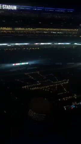 A stadium with the words "METLIFE STADIUM" in the upper left corner is illuminated with bright lights.  A large crowd fills the stands, with a formation of performers holding lights visible on the field spelling out "SNL" at the center. A person wearing a Giants hat stands in the foreground, partially obstructing the view of the field.