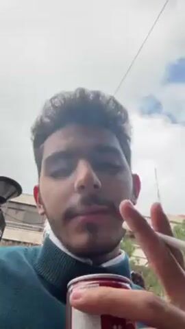 A young man is shown at a low angle, holding a lit cigarette and a can of Red Berry Ice Tea. He takes a drag from the cigarette and smoke is exhaled in front of his face. He is wearing a green turtleneck sweater over a collared shirt. The background shows a cloudy sky, buildings and trees.