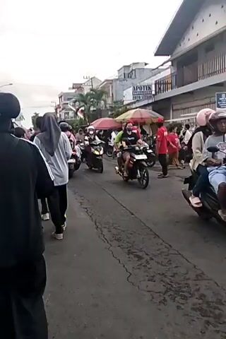 The video shows a street scene, likely in Southeast Asia, with numerous motorcycles and pedestrians. The road is paved and appears to have some cracks and wear. Buildings line the street, some with signs that are partially visible, including one for "APOTEK". There are market stalls with colorful umbrellas set up along the side of the road. Many people are on motorcycles, some riding alone and others with passengers. Pedestrians are walking on the side of the road. The overall impression is a busy, everyday scene.