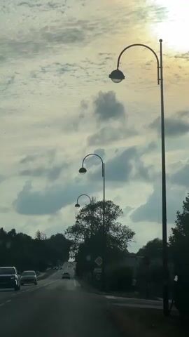 The video captures a view from a vehicle moving along a road, likely in the daytime, given the bright sky and sunlight. The sky is filled with clouds, some appearing dark and dramatic, while others are lighter and more diffused. A prominent sunbeam is visible in the upper right corner of the frame. Along the right side of the road, there are several streetlights with curved arms, spaced at intervals. One streetlight is closer to the camera and appears larger than the others. There is also a large tree with dense foliage next to the road. Traffic signs are visible, including a diamond-shaped sign with a yellow center and a circular sign with the number 70, indicating a speed limit. A few cars are driving on the road ahead, and some appear to be moving in the opposite direction. The road itself has clear lane markings, with white dashed lines separating the lanes. The environment appears to be somewhat rural or suburban, with trees and vegetation lining the sides of the road. The overall impression is of a journey or drive through a scenic, possibly overcast, landscape.