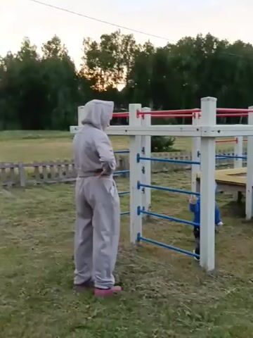 This video captures a moment on an outdoor playground, likely in Smolino, Russia, as indicated by the context. A woman, dressed in a light grey tracksuit with a hood and pink footwear, stands on the left side of the frame, facing a young child. The child, wearing a blue jacket and a grey hat, is positioned within a white and blue jungle gym structure. The playground equipment features white vertical posts and blue horizontal bars, with red accents on some of the upper rails.

In the background, a colorful playhouse with a yellow, red, and blue roof is visible, along with more playground structures and a yellow slide. The setting is a grassy area with scattered dry leaves and some patchy bare ground, suggesting it might be late summer or early autumn. A line of trees forms the backdrop, with the sky above appearing overcast or at dusk, indicating it's likely evening.

The woman appears to be observing the child, her hands on her hips. The child is looking towards the woman and the jungle gym, seemingly engaged with the play structure. The overall mood appears calm and relaxed, suggesting a casual outing to the playground. There is no visible text in the video.