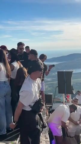 A group of people are gathered on a raised platform or balcony, possibly a rooftop or mountaintop, overlooking a body of water and a distant landscape. The sky is blue with some clouds. The people are dressed in casual summer attire, including t-shirts, shorts, and hats. Some are holding drinks, and one person is smoking. A speaker is visible on the right side of the frame, suggesting music is playing or about to play. The overall atmosphere appears to be a social gathering or outdoor party.