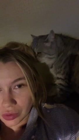 A young woman with light brown hair and fair skin is recording a selfie video. She is wearing a blue and white checkered shirt. She is looking directly at the camera with a neutral expression. Behind her is a grey tabby cat that is sitting very close to her head. The woman says "че моя девочка", which translates to "what is my girl doing?". She then says "Ой, ты что толкаешься", which translates to "Oh, what are you pushing?". The background is a light beige wall. The lighting is slightly dim.