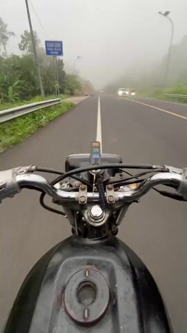 The video captures a first-person perspective of someone riding a motorcycle down a two-lane asphalt road in Karangmangu, Indonesia. The scene is dominated by heavy fog, significantly reducing visibility ahead.

The motorcycle's handlebars, fuel tank, and dashboard area are visible in the foreground, showing a worn black tank and chrome handlebars. The rider is moving forward on the right side of the road, which has a solid white line dividing the two directions of traffic and yellow lines marking the shoulder.

Several other vehicles are on the road: a black pickup truck is seen moving away in the distance on the right, and other cars and trucks are dimly visible through the fog further ahead. Lush green trees and vegetation line the left side of the road, and a street light pole is also present. A blue road sign with illegible white text can be seen on the left side of the road at the beginning of the video. The weather is overcast and foggy, creating a soft, diffused light.