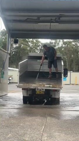 A man is standing in the bed of a white dump truck and using a pressure washer to clean the inside of the truck bed. The truck is parked under a covered structure, and the ground is wet, indicating that a washing station is in use. The man is wearing a grey t-shirt, dark shorts, and work boots. He is holding a pressure washer wand with a blue hose connected to it. The truck has a yellow license plate with the characters DJ 84 TC. Trees and buildings are visible in the background.
